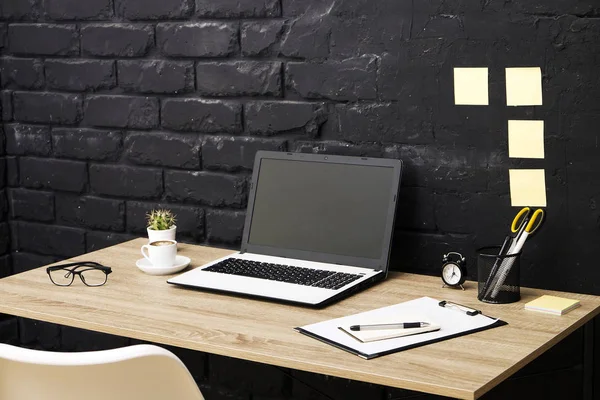 Creative workspace of a blogger. White laptop computer & folded eye glasses on wooden table in loft style office with black brick walls. Designer's table concept. Close up, copy space, background.