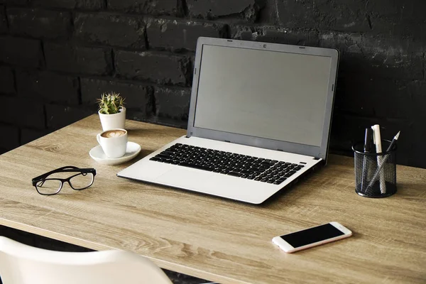 Creative workspace of a blogger. White laptop computer & folded eye glasses on wooden table in loft style office with black brick walls. Designer\'s table concept. Close up, copy space, background.