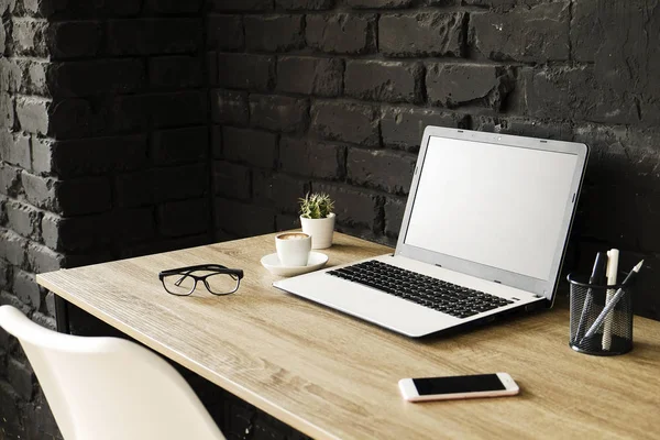 Creative workspace of a blogger. White laptop computer & folded eye glasses on wooden table in loft style office with black brick walls. Designer\'s table concept. Close up, copy space, background.