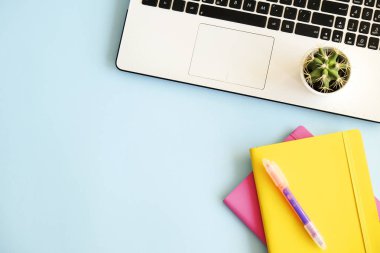 Ofis İşyeri dizüstü bilgisayar, birden çok renkli Notebook, yeşil kaktüs bitki, kahve, sarf malzemeleri ve kırtasiye mavi zemin üzerine beyaz fincan ile. Yakın çekim, kopya alanı, üstten görünüm, düz lay.