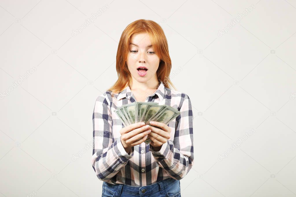 Young beautiful redhead woman screaming in rejoice, laughing w/ open mouth, fistful of one hundred dollar bills like fan. Excited attractive female with lots of cash. Background, copy space, close up.