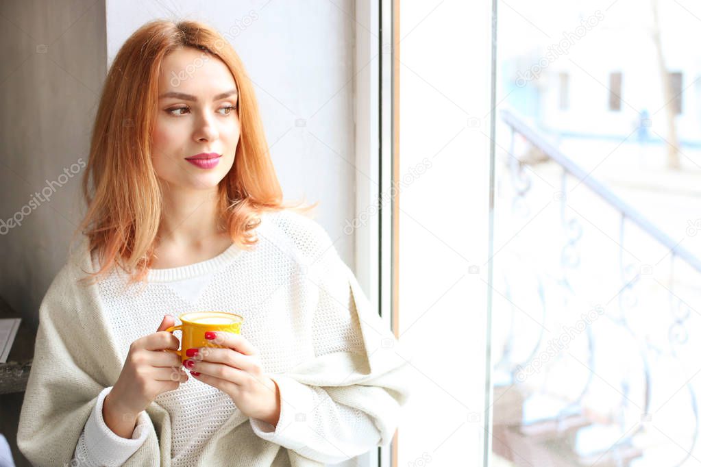 Beautiful redhead woman, sitting near coffee shop window holding yellow cup of hot beverage, wearing winter autumn season white knitted sweater & big loose scarf on shoulders. Background, close up.