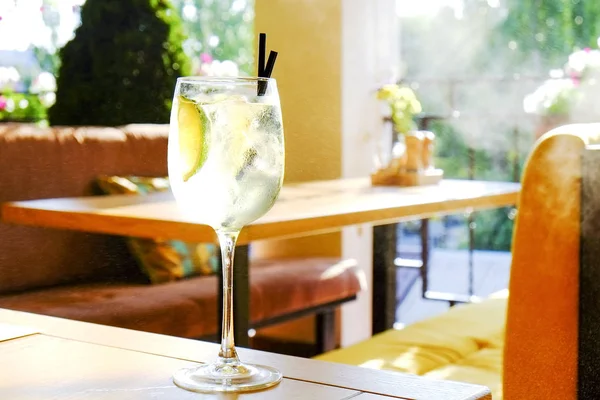 Glass of fresh summer non alcoholic refreshing cocktail with ice & lemon, standing on table of patio outside hip restaurant with city greenery view. Soft sunlight ray. Background, copy space, close up