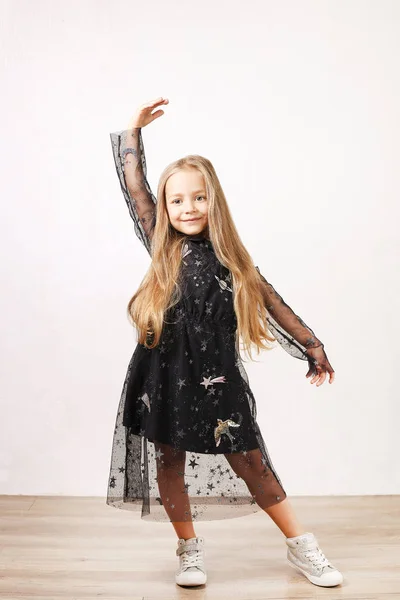 Little blonde girl with long golden hair wearing black dress with stars & planets, dancing, smling & having fun, white wall background. Five years old blonde female child posing. Copy space, close up.