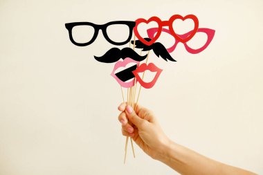 Çubuk kırmızı kağıttan yapılmış üzerinde kalp şeklinde karnaval Aksesuarları, dudaklar ve gözlük tutan genç kadın el kapatın. Erkek parti seti konsepti ile. Sarı duvar arka plan, kopya alanı.