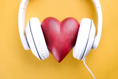 Senin kalp kavramı dinle. Parlak sarı zemin üzerinde beyaz kulaklık ile büyük kırmızı kalp. Sevgililer günü kompozisyon kulaklık ve kalp. Yakın çekim, kopya alanı, üstten görünüm, düz lay,
