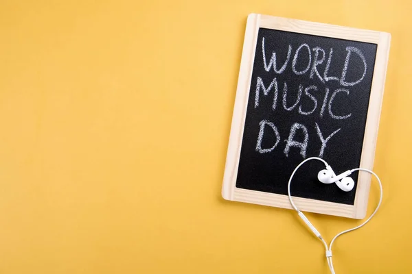 White earphones with wooden blackboard with world music day text written on it. Minimalistic composition with earbuds & chalkboard on juicy yellow background. Close up, top view, copy space, flat lay