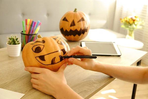 Woman\'s hands drawing scary and funny Jack O Lantern face on ripe orange pumpkin with black marker pen for all hallow eve Halloween party. Holiday decoration concept. Close up, copy space, background.