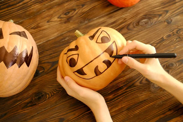 Woman\'s hands drawing scary and funny Jack O Lantern face on ripe orange pumpkin with black marker pen for all hallow eve Halloween party. Holiday decoration concept. Close up, copy space, background.