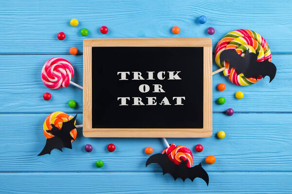 Trick or treat text written on blackboard with decorative paper bats, eyeball shaped candy, gummy worms, spiders & bones. Halloween decor concept. Background, copy space, close up, top view, flat lay.