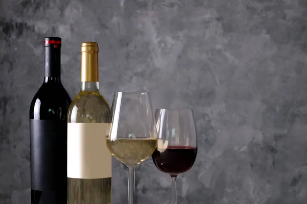 Two bottles of red and white wine with matte black & white blank labels on concrete wall background. Wineglasses half full with vintage expensive wine of different type. Close up, copy space.