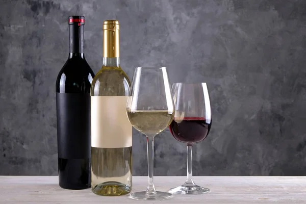 Two bottles of red and white wine with matte black & white blank labels on concrete wall background. Wineglasses half full with vintage expensive wine of different type. Close up, copy space.