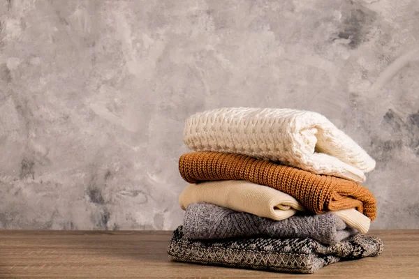 Bunch of knitted warm pastel color sweaters, different knitting patterns folded in messy stack on brown wooden table, concrete texture wall background. Fall winter season knitwear. Close up, copy space