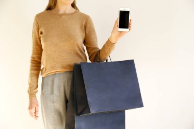 Genç kadın klasik şık kıyafet, kahverengi süveter içinde ince ve gevşek gri pantolon alışveriş gitti, kağıt torba tutarak ve boş ekran smartphone gösterilen. Beyaz arka plan, kopya alanı, yakın çekim.