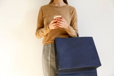 Genç kadın klasik şık kıyafet, kahverengi süveter içinde ince ve gevşek gri pantolon alışveriş gitti, kağıt torba tutarak ve Smartphone'da sohbet. Beyaz arka plan, kopya alanı, yakın çekim.