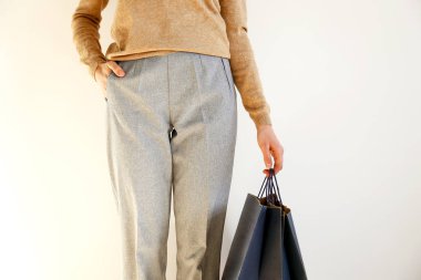 Genç kadın klasik şık kıyafeti slim, kahverengi süveter ve gevşek gri pantolon cebinde, alışveriş, kağıt torba tutarak el. Beyaz arka plan, kopya alanı, yakın çekim.