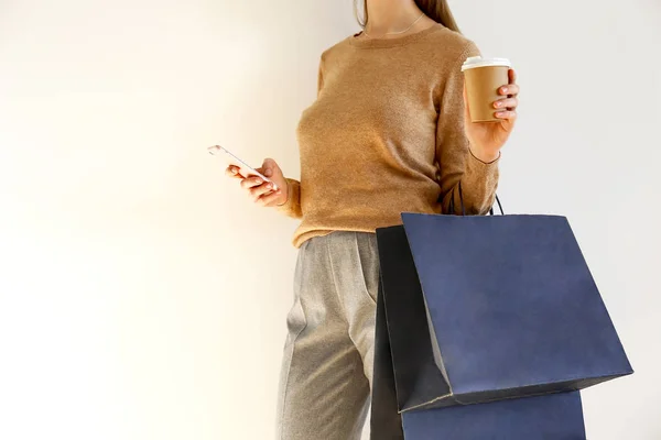 年轻苗条的妇女在经典时尚的服装 棕色毛衣和宽松的灰色裤子去购物 拿着纸袋 在智能手机上聊天 喝咖啡 背景白色 复制空间 — 图库照片