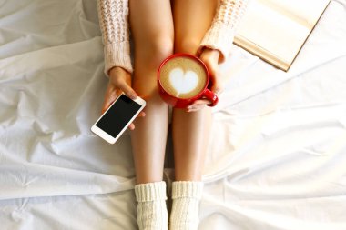 Tembel öğleden sonra kavram. Yatakta, oturan kadın kırmızı fincan cappuccino kahve latte sanat köpük ve boş ekran cep telefonu şeklinde kalp ile tutarak kitap, okuma görüntüsünü kapatın. Arka plan, kopya alanı.
