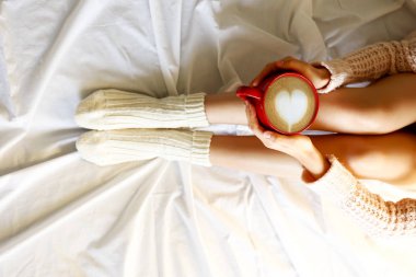 Cappuccino kahve içme ve beyaz harmanlamıştır ile yatakta oturan genç kadın. Üstten görünüm sıcak yün çorap ve kazak içinde kadın bacak. Konfor kış tatil kavramı. Yakın çekim, uzay, arka plan kopya.