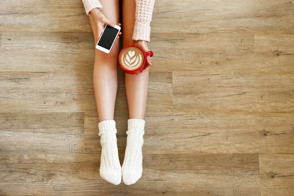 Lazy afternoon concept. Young woman wearing knee high socks, knitted sweater, sitting on wooden floor, holding blank screen mobile phone, red cup of coffee. Background, copy space, top view, close up.