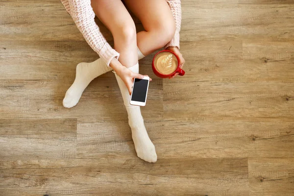 Lazy afternoon concept. Young woman wearing knee high socks, knitted sweater, sitting on wooden floor, holding blank screen mobile phone, red cup of coffee. Background, copy space, top view, close up.