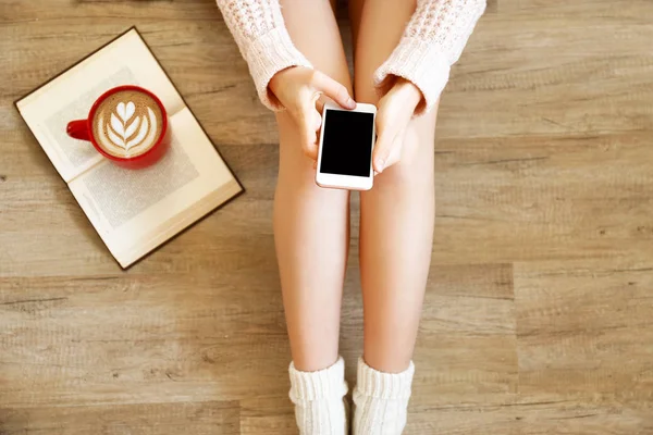 Lazy afternoon concept. Young woman wearing knee high socks, knitted sweater, sitting on wooden floor, blank screen mobile phone, red cup of coffee, book. Background, copy space, top view, close up.