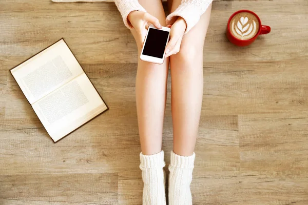 Lazy afternoon concept. Young woman wearing knee high socks, knitted sweater, sitting on wooden floor, blank screen mobile phone, red cup of coffee, book. Background, copy space, top view, close up.
