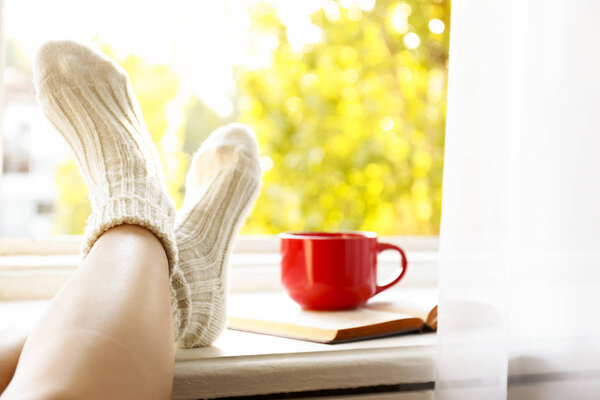 Young woman sitting with crossed legs in warm woolen socks leaning on windowsill, cup of coffee and book, autumnal window view w/ yellow leaves. November mood concept. Background, copy space, close up