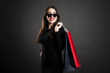 Siyah Cuma satış konsepti. Gülümseyerek, seksi elbise ve kedi göz güneş gözlüğü, boş alışveriş torbaları siyah arka plan üzerinde tutarak takıyor çekici genç kadın uzun esmer saçlı. Uzay kopyalayın, yakın çekim.