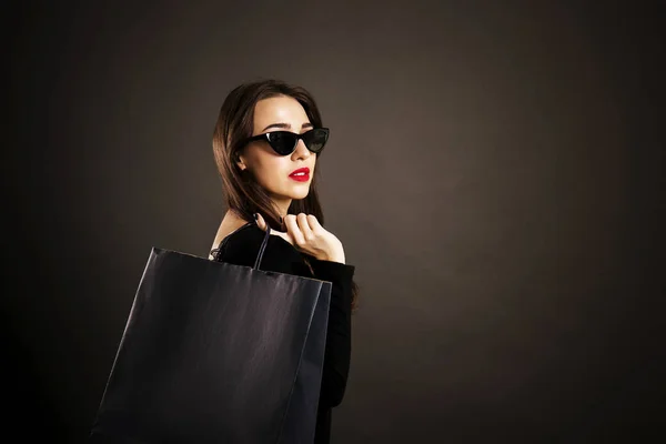 黑色星期五销售概念 有吸引力的年轻妇女与长黑发头发 穿着性感的礼服和猫眼太阳眼镜 持有空白购物袋在黑色背景 复制空间 — 图库照片