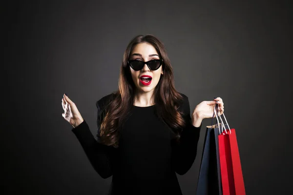 Black friday sale concept. Attractive young woman with long brunette hair, smiling, wearing sexy dress & cat eye sunglasses, holding blank shopping bags over black background. Copy space, close up.