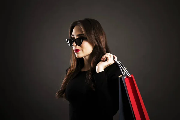 黑色星期五销售概念 有吸引力的年轻妇女与长黑发头发 穿着性感的礼服和猫眼太阳眼镜 持有空白购物袋在黑色背景 复制空间 — 图库照片