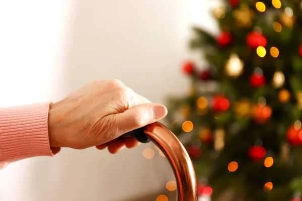 Elderly woman celebrating Christmas at home, with decorated holiday pine tree on background. Old lady at nursing home. Close up, copy space, cropped shot.