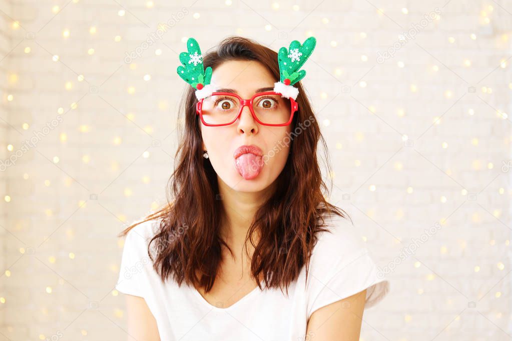 Beautiful young woman wearing santa claus furry hat & masquerade glasses for Chritmas celebration event. Attractive female being silly, making faces at dress up party. Background, close up, copy space