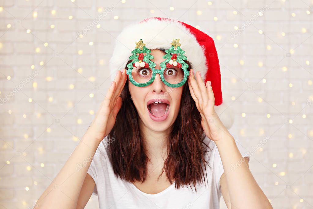 Beautiful young woman wearing santa claus furry hat & masquerade glasses for Chritmas celebration event. Attractive female being silly, making faces at dress up party. Background, close up, copy space