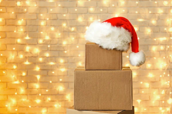 Blank brown freight box with Santa Claus hat on top, brick wall with Christmas lights on background. Moving company / delivery service holiday deals promotion concept. Copy space, close up.