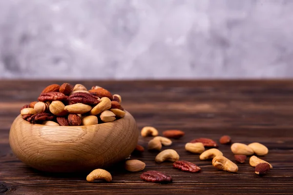 Mixed nuts in wooden bowl and scattered on table. Trail mix of pecan, almond, macadamia & brazil edible nuts with walnut hazelnut on wood textured surface. Background, copy space, top view, close up.