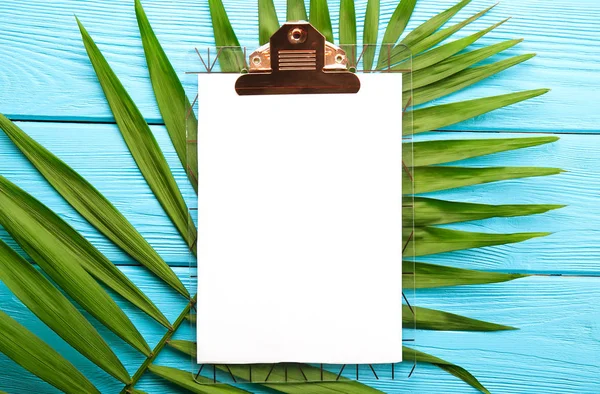 Clipboard with blank piece of paper on big green leaf of parlor palm tree, blue wooden textured table background. Empty sheet on branch of tropical plant. Top view, close up, copy space, flat lay.