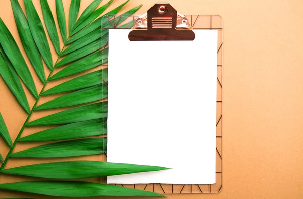 Clipboard with blank piece of paper on big green leaf of parlor palm tree, golden orange gradient table background. Empty sheet on branch of tropical plant. Top view, close up, copy space, flat lay.