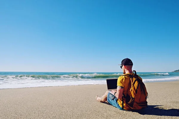 Traveler writing blog entry on white laptop, sitting at exotic empty beach. Freelance remote work concept. Self employed fit young male in bright yellow t-shirt coding. Copy space, sea view background