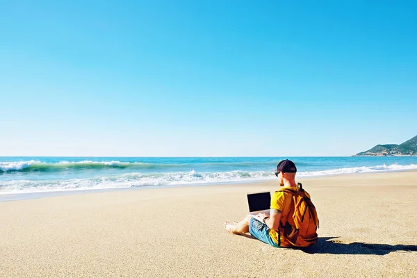 Traveler writing blog entry on white laptop, sitting at exotic empty beach. Freelance remote work concept. Self employed fit young male in bright yellow t-shirt coding. Copy space, sea view background