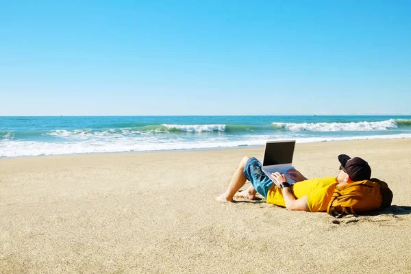 Traveler writing blog entry on white laptop, lying at exotic empty beach. Freelance remote work concept. Self employed fit young male in bright yellow t-shirt coding. Copy space, sea view background