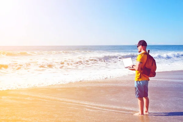 Fit travel blogger holding laptop, writing article on white laptop, standing on empty beach. Freelance remote work concept. Self employed man in yellow t-shirt coding. Copy space, sea view background.
