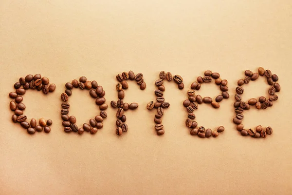 Palavra de café soletrado com grãos torrados sobre fundo marrom cremoso. Close-up, espaço de cópia, vista superior, flat lay . — Fotografia de Stock