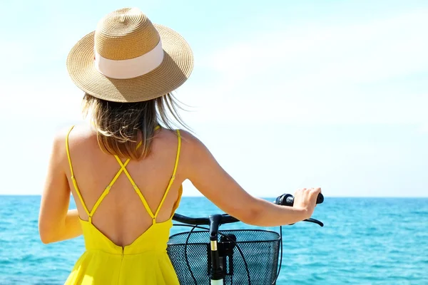 Ung bekymmerslös kvinna i ljust gul klänning med cykel vid havet stranden. Oigenkännlig kvinna bär bred brätte hatt cykling på sandstrand på solig dag. Kopiera utrymme, bakgrund. — Stockfoto