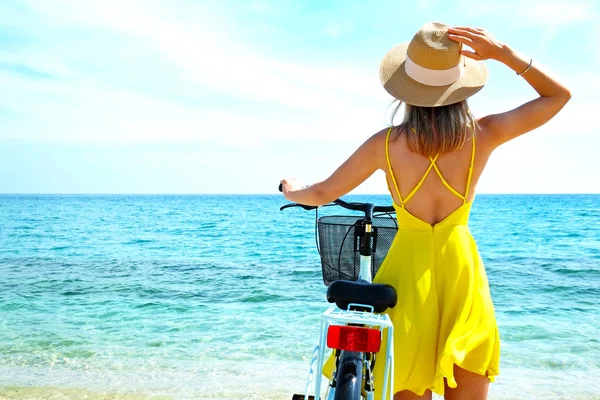 Young carefree woman in bright yellow dress with bicycle at ocean beach. Unrecognizable female wearing broad brim straw hat biking on sandy sea shore on sunny day. Copy space, background. — ストック写真