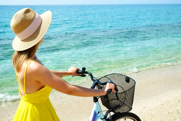 Young carefree woman in bright yellow dress with bicycle at ocean beach. Unrecognizable female wearing broad brim straw hat biking on sandy sea shore on sunny day. Copy space, background. — ストック写真