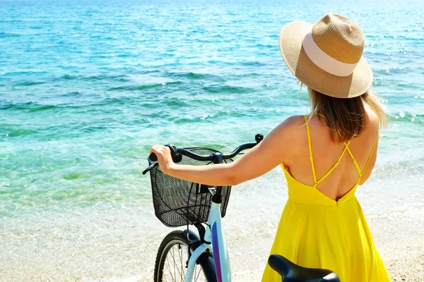 Young carefree woman in bright yellow dress with bicycle at ocean beach. Unrecognizable female wearing broad brim straw hat biking on sandy sea shore on sunny day. Copy space, background. — ストック写真