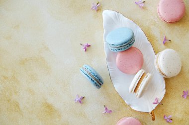 Farklı renk ve tat fransız makarna tatlılar demet minimalist kompozisyon, leylak çiçekli sarı beton doku arka plan üzerinde dağınık. Üstten görünüm, yakın çekim, düz döşeme, kopyalama alanı.