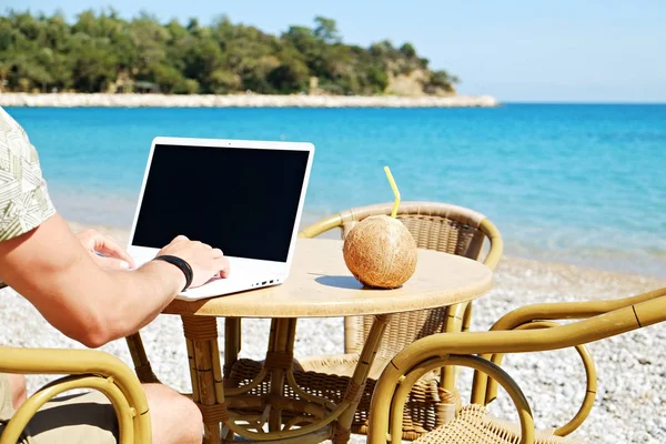 Young man working on his laptop at the beach at tropical destination. Office on the beach concept.